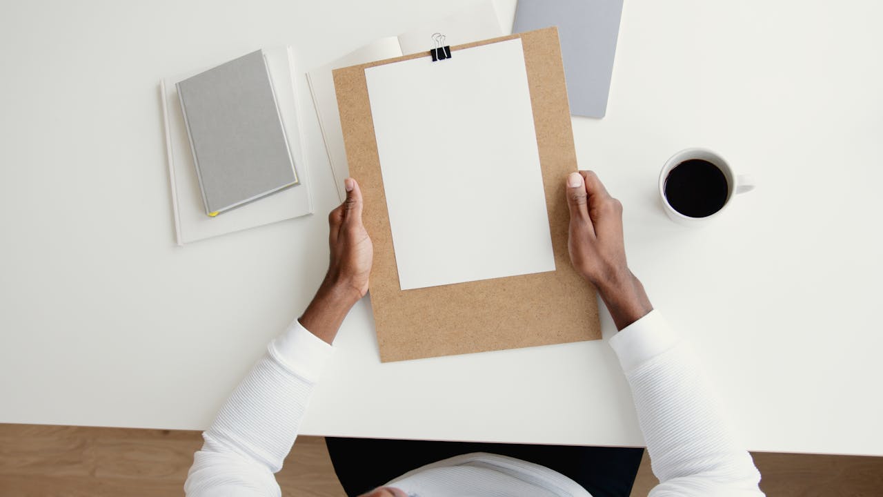 Top view of a person holding a clipboard with blank paper on a desk with coffee and books.